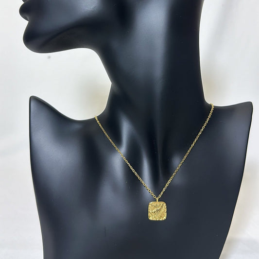 A smooth black mannequin model wearing a gold chain necklace with a statement square pendant featuring raised decorative details.