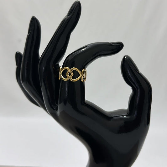 A delicate gold ring with a series of interconnected heart motifs is displayed on a black mannequin hand with fingers slightly curled. The background is smooth and white, giving the ring a striking contrast.