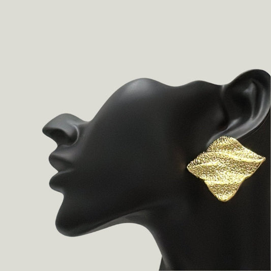 Side profile of a black matte mannequin head wearing a large, textured gold earring shaped like a leaf , set against a plain white background.