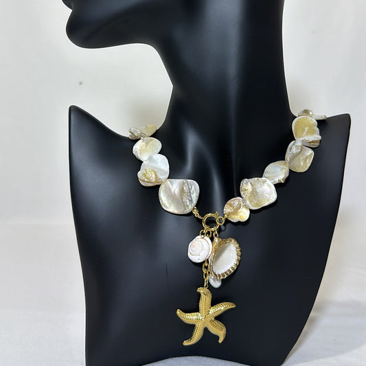 A necklace with large, shiny shell-like beads in off-white and light gold hues, complemented by a dangling gold starfish and seashell charms, is showcased on a black matte mannequin bust with a minimalistic white backdrop.