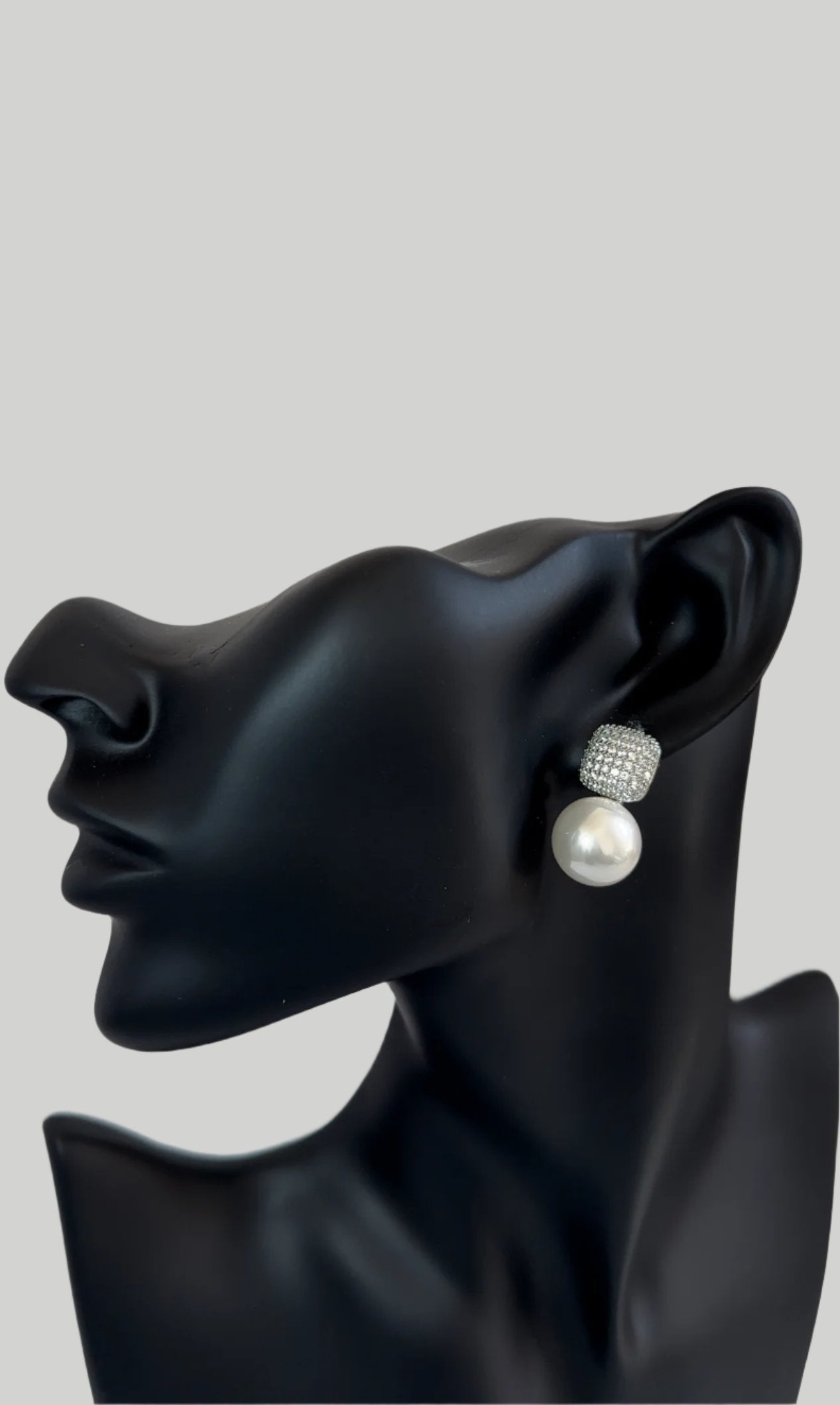  A mannequin head with a sleek black finish wearing a stud earring featuring a large pearl drop and a square, diamond-encrusted setting at the top. The background is neutral, allowing focus on the earring's elegant design.