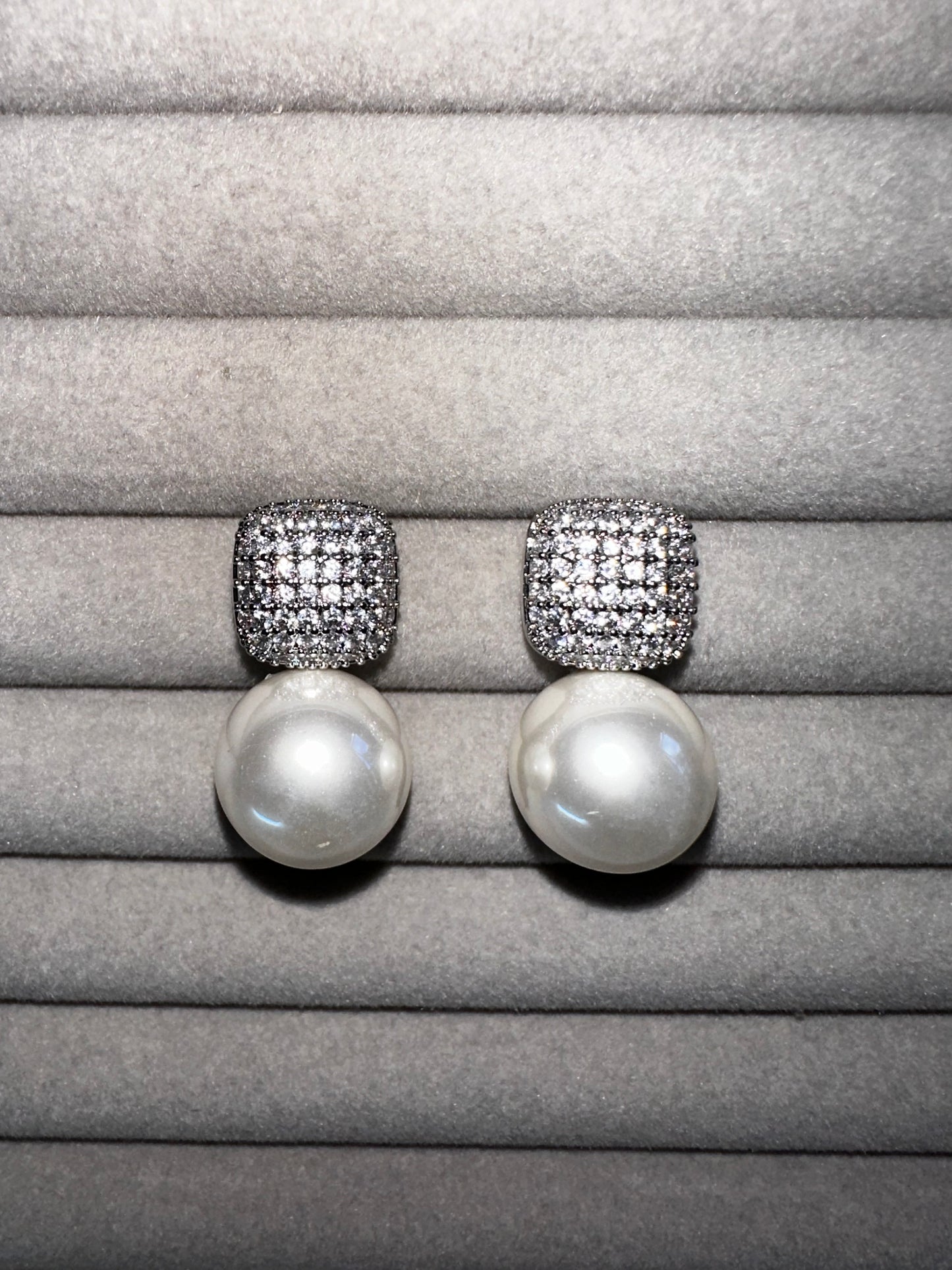 A pair of pearl drop earrings with square, diamond-encrusted tops displayed on a soft, gray velvet surface. The pearls are large and round, contrasting with the sparkle of the diamonds.