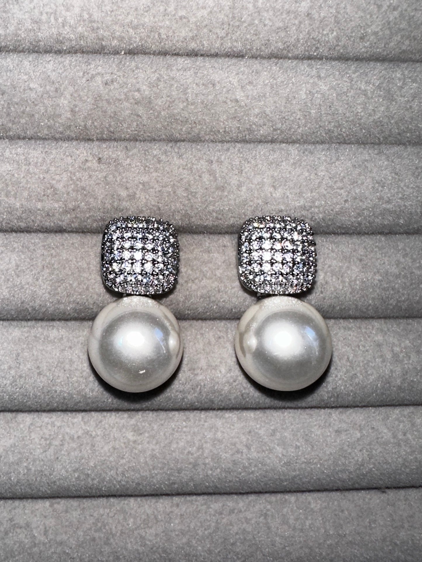 A pair of pearl drop earrings with square, diamond-encrusted tops displayed on a soft, gray velvet surface. The pearls are large and round, contrasting with the sparkle of the diamonds.