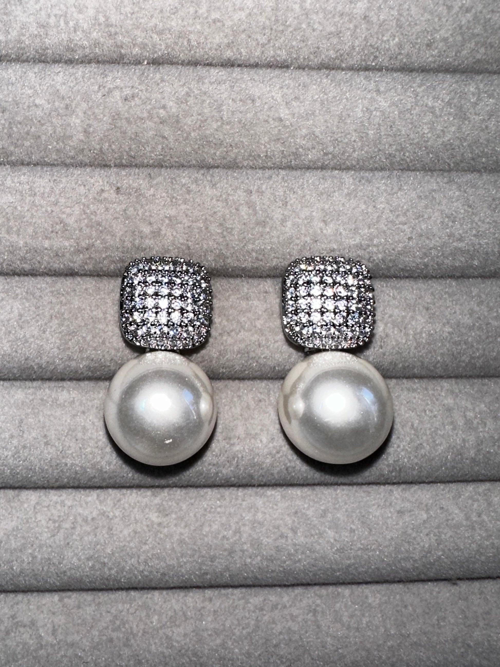 A pair of pearl drop earrings with square, diamond-encrusted tops displayed on a soft, gray velvet surface. The pearls are large and round, contrasting with the sparkle of the diamonds.