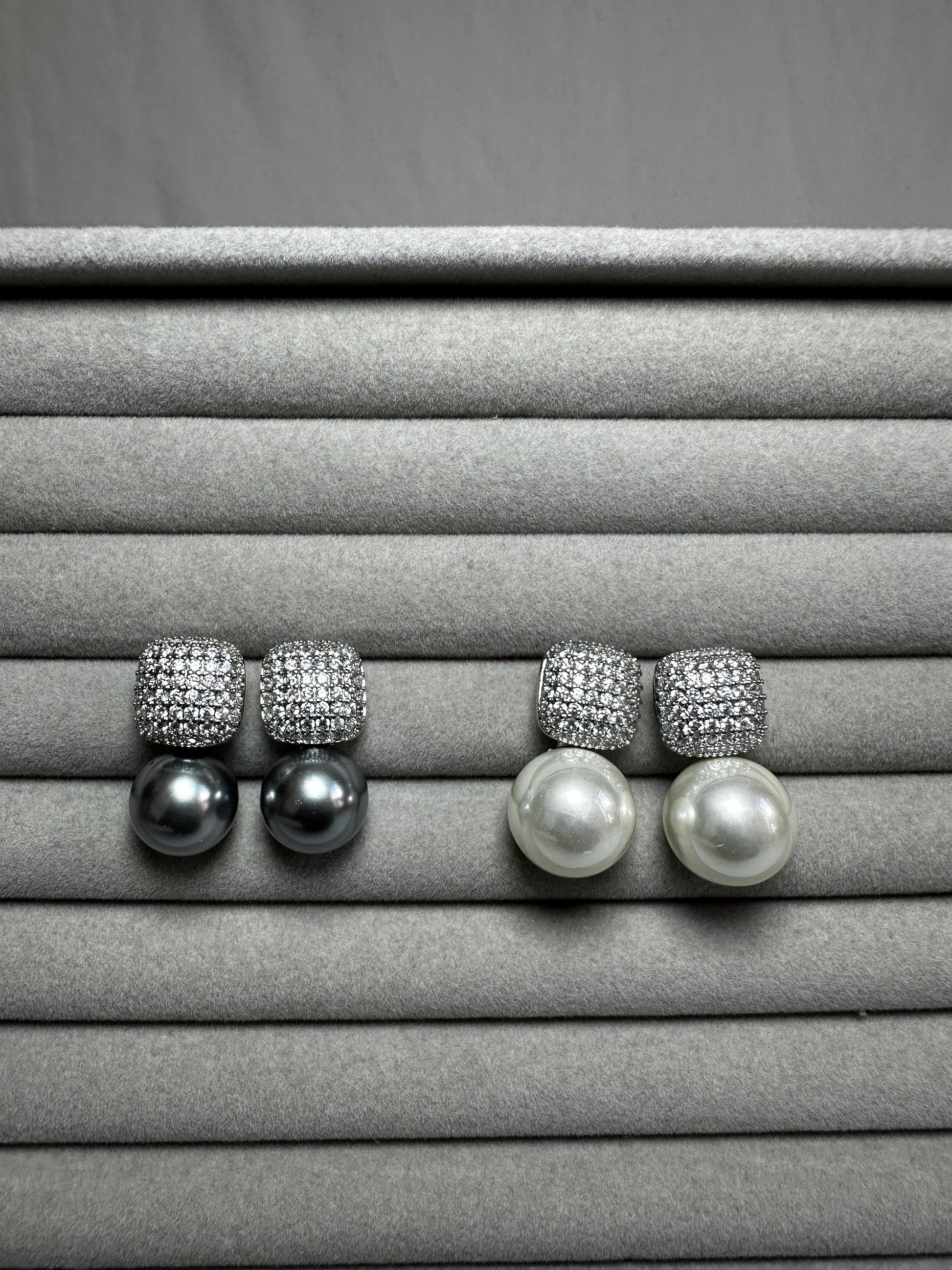 Two pairs of pearl drop earrings with square, diamond-encrusted tops displayed on a soft, gray velvet surface. The pearls are large and round, contrasting with the sparkle of the diamonds.