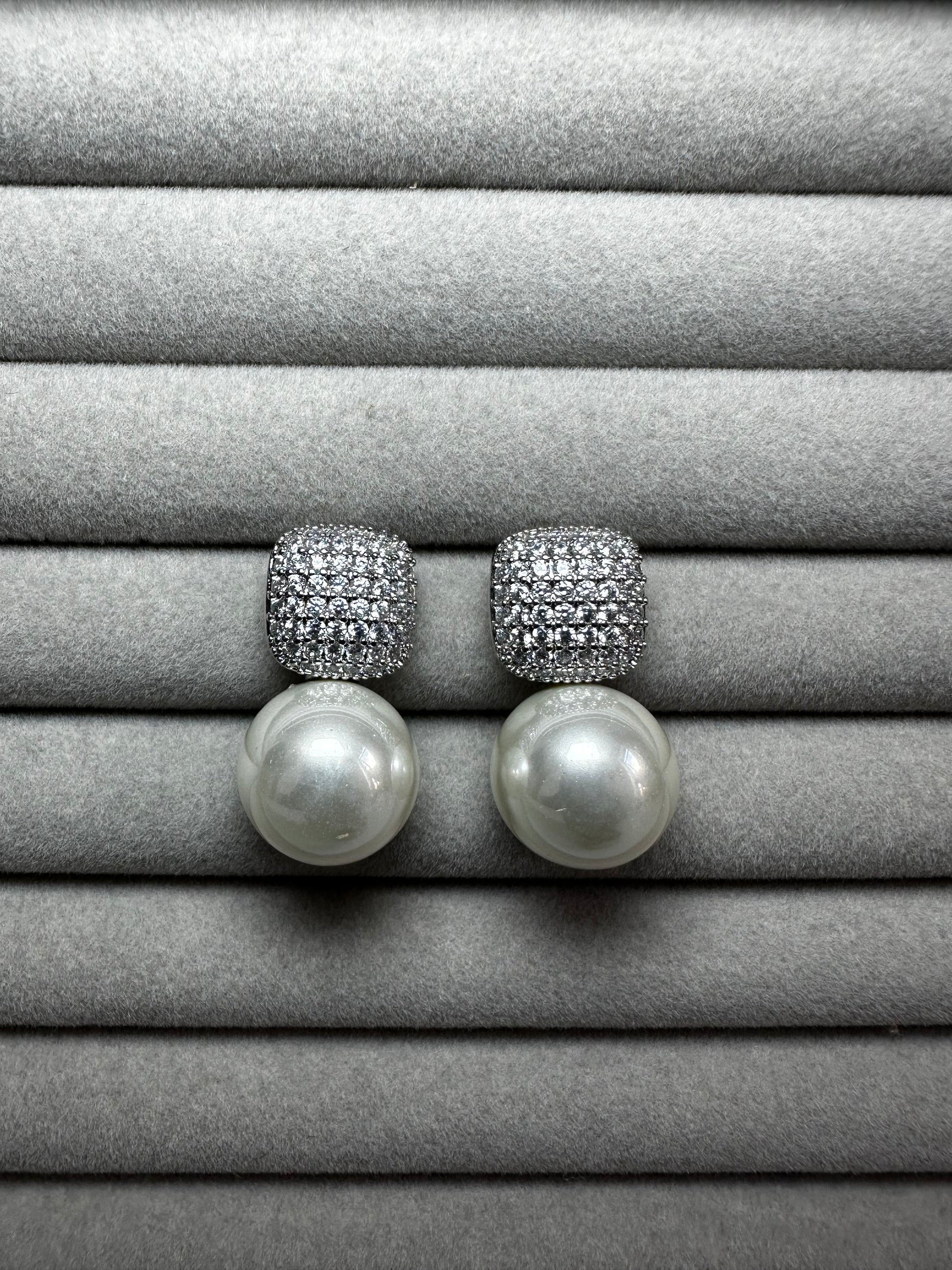 A pair of pearl drop earrings with square, diamond-encrusted tops displayed on a soft, gray velvet surface. The pearls are large and round, contrasting with the sparkle of the diamonds.