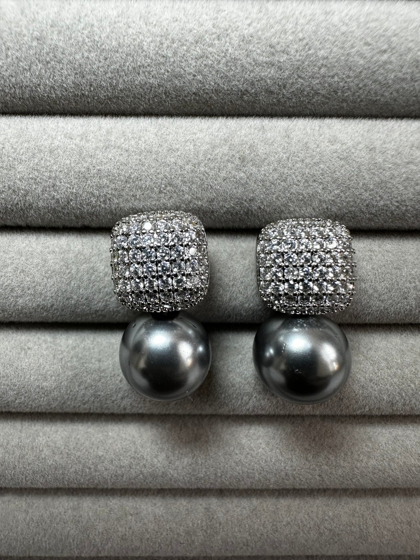 A pair of pearl drop earrings with square, diamond-encrusted tops displayed on a soft, gray velvet surface. The pearls are large and round, contrasting with the sparkle of the diamonds.