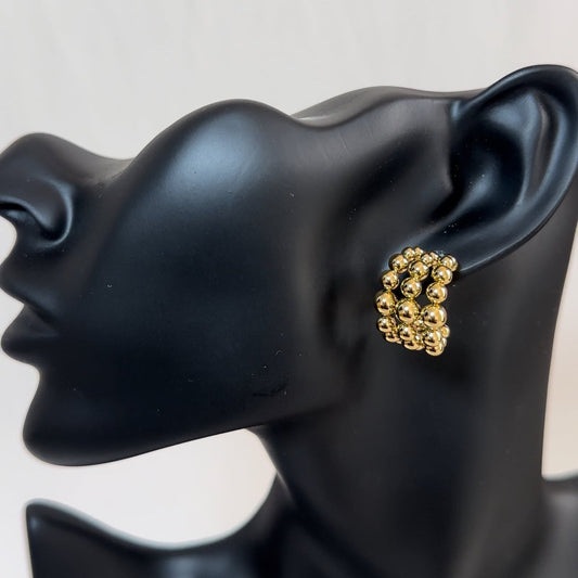 Profile of a sleek, black mannequin wearing a chunky gold earring composed of several overlapping metallic beads.